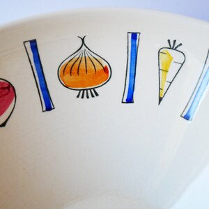 May include: White ceramic bowl with a colorful vegetable design. The bowl features red, orange, yellow, blue, and black painted vegetables, including a carrot, onion, and beets.