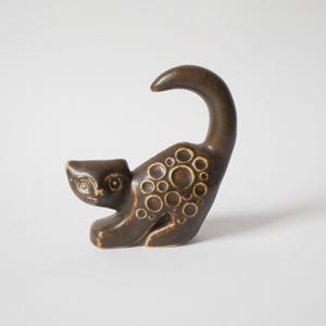 Puede incluir: Figura de gato de cerámica marrón con cola curvada y patrones circulares. El gato está en una pose juguetona, con la cabeza inclinada. Un artículo decorativo para el hogar.