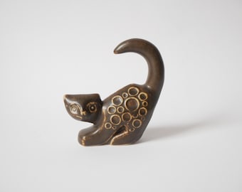 Może przedstawiać: Brązowa ceramiczna figurka kota z zakrzywionym ogonem i okrągłymi wzorami. Kot jest w zabawnej pozie, z pochyloną głową. Dekoracyjny przedmiot do domu.