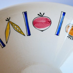 May include: White ceramic bowl with a colorful vegetable design. The bowl features a red tomato, yellow pea pods, a blue onion, and a green leaf.