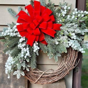 May include: A Christmas wreath made of grapevine with frosted pine branches and white berries. A large red velvet bow is centred on the wreath.