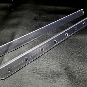 K&ouml;nnte beinhalten: Zwei klare, starre Acrylstreifen. Ein Streifen ist glatt mit einer geraden Kante. Der zweite Streifen weist eine Reihe von gleichm&auml;&szlig;ig verteilten, kreisf&ouml;rmigen L&ouml;chern auf, die entlang der L&auml;nge zur Befestigung eingestanzt sind.