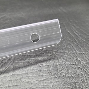 K&ouml;nnte beinhalten: Ein durchsichtiger, flexibler Kunststoffstreifen mit einem glatten, l&auml;nglichen Design und einem einzelnen kreisf&ouml;rmigen Loch nahe dem Ende. Das Material ist transparent und halbstarre, mit einer Reihe feiner, paralleler Einkerbungen.