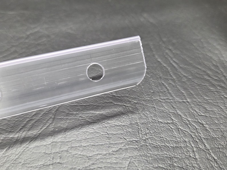 K&ouml;nnte beinhalten: Ein durchsichtiger, flexibler Kunststoffstreifen mit einem glatten, l&auml;nglichen Design und einem einzelnen kreisf&ouml;rmigen Loch nahe dem Ende. Das Material ist transparent und halbstarre, mit einer Reihe feiner, paralleler Einkerbungen.
