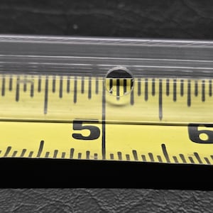K&ouml;nnte beinhalten: Ein klarer, transparenter Kunststoffstreifen mit einem kleinen, kreisf&ouml;rmigen Loch, das direkt &uuml;ber der 12,7-cm-Markierung eines gelben Ma&szlig;bandes positioniert ist. Das Band zeigt schwarze Ziffern und Skalenstriche f&uuml;r pr&auml;zise Messungen.