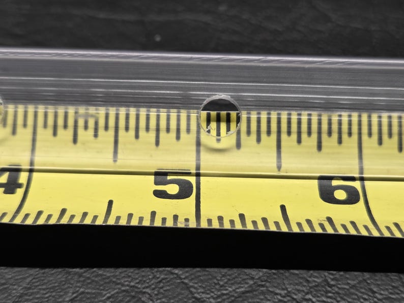 K&ouml;nnte beinhalten: Ein klarer, transparenter Kunststoffstreifen mit einem kleinen, kreisf&ouml;rmigen Loch, das direkt &uuml;ber der 12,7-cm-Markierung eines gelben Ma&szlig;bandes positioniert ist. Das Band zeigt schwarze Ziffern und Skalenstriche f&uuml;r pr&auml;zise Messungen.