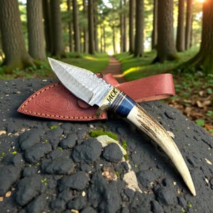 May include: A handcrafted hunting knife with a patterned steel blade and a handle made of bone and wood. The knife is resting on a brown leather sheath with the name "JACK" engraved on the blade. The background is a forest scene.