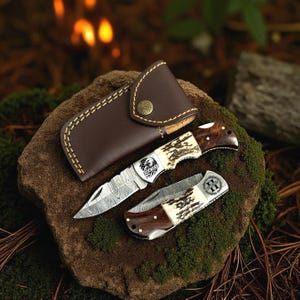 May include: A set of two folding knives with a brown leather sheath. The knives have patterned blades, antler-style handles, and metal accents. One knife features an eagle design and the text "YOUR TEXT". The sheath is brown with a button closure.