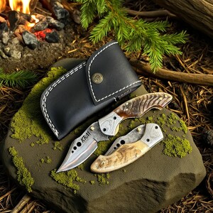 May include: Two folding knives with patterned blades and wooden handles rest on a mossy rock, next to a black leather sheath. The knives have the initials "KRM" etched on the blade. The background includes a campfire and pine needles.