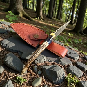 May include: A handcrafted hunting knife with a patterned blade and a handle made of antler. The knife is accompanied by a brown leather sheath with embossed detailing and a brass button closure. The knife is displayed outdoors on a bed of rocks and foliage.