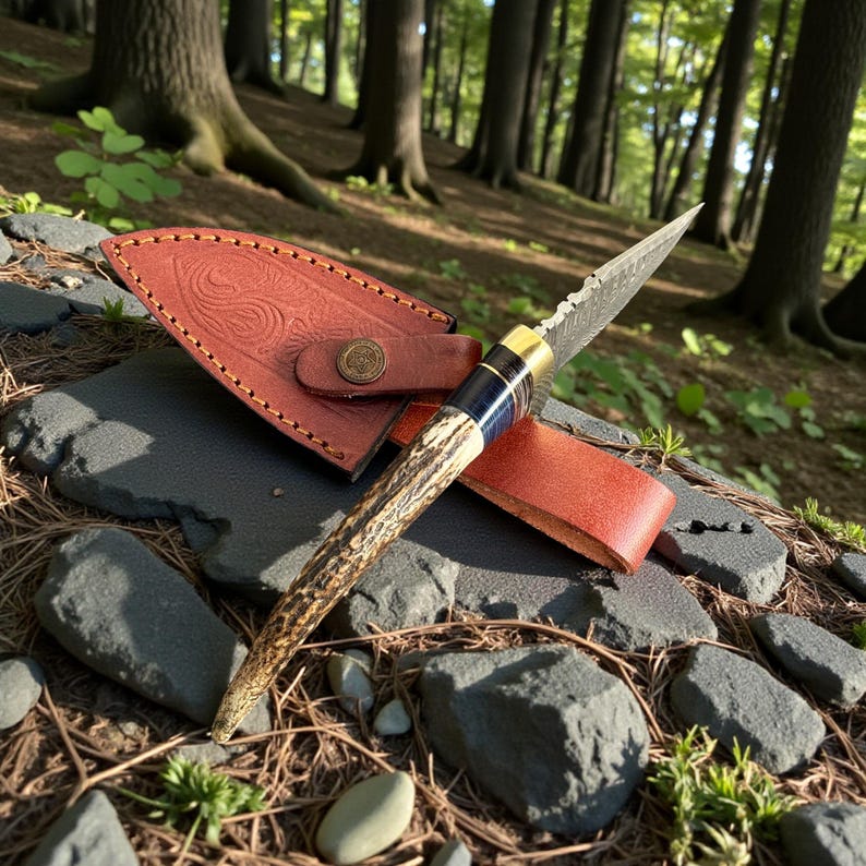 May include: A handcrafted hunting knife with a patterned blade and a handle made of antler. The knife is accompanied by a brown leather sheath with embossed detailing and a brass button closure. The knife is displayed outdoors on a bed of rocks and foliage.
