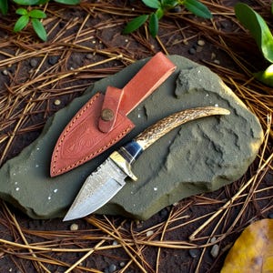 May include: A handcrafted hunting knife with a patterned blade and an antler handle. The knife rests on a gray stone next to a brown leather sheath. The sheath has a decorative embossed design and a snap closure.