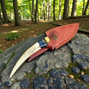 May include: A handcrafted knife with a bone handle and a leather sheath. The knife features a curved blade and a handle wrapped with blue and black twine. The sheath is brown with an embossed floral design and a button closure.