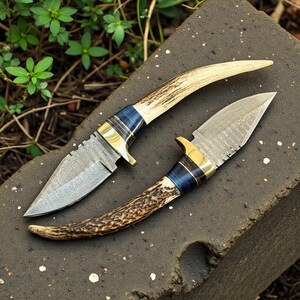 May include: Two handmade knives with patterned steel blades and antler handles. The handles feature blue and gold accents. The blades have a pointed tip and a decorative pattern. The knives are displayed on a dark gray surface.