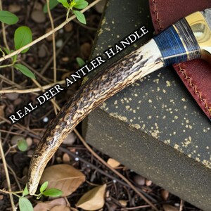 May include: Close-up of a knife handle made from a real deer antler. The handle has a natural, textured appearance with shades of brown and cream. The text "REAL DEER ANTLER HANDLE" is printed on the image.