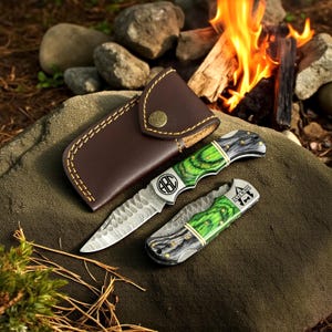 May include: Two knives with green and black handles and a brown leather sheath. One knife is open, displaying a serrated blade and the initials "ABC". The other knife is closed, with the text "YOUR TEXT" visible. The sheath is closed with a brass button.