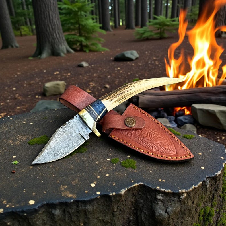 May include: A Damascus steel knife with a curved antler handle and a brown leather sheath. The knife is resting on a dark, textured surface, with a campfire in the background. The blade has a unique pattern.