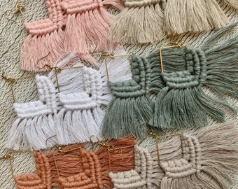 May include: A set of six pairs of handmade macrame earrings with fringe. The earrings are made with natural fibers in a variety of colors, including beige, white, pink, green, and brown. Each pair features a square shape with a gold hoop.