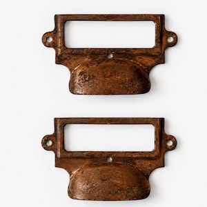 May include: Two rectangular metal drawer pulls with a dark, weathered, rusted brown finish. Each pull features a central label holder frame with an open rectangular centre and a curved handle grip below, including two side screw holes for mounting.