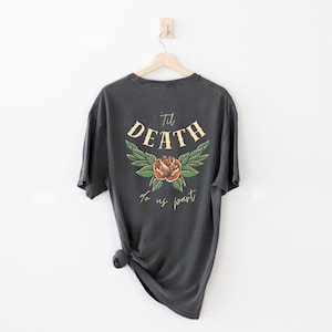 May include: Dark gray t-shirt with a floral design and the text "Til Death To Us Part". The design features a red rose with green leaves and the word "DEATH" in a yellow, arched font. The shirt is hanging on a wooden hanger.