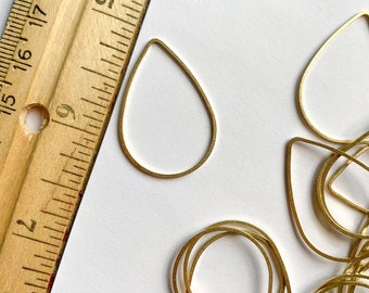 Puede incluir: Primer plano de un montón de blanks de pendientes en forma de lágrima de metal dorado. Los blanks están dispuestos en un patrón aleatorio sobre una superficie blanca. Una regla es visible en el fondo.