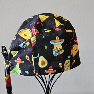 May include: Black surgical scrub cap with a colorful pattern of cartoon tacos, avocados, and musical characters wearing sombreros. The cap has a tie closure for an adjustable fit.