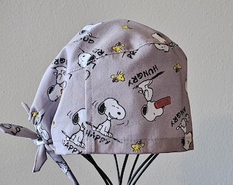May include: A light purple surgical scrub cap featuring a repeating pattern of Snoopy characters in various poses, along with the words "Happy", "Hungry", and "Angry". The cap has a tie closure and is displayed on a black stand.