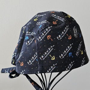 May include: Black scrub hat with a repeating pattern of the Friends logo and colorful umbrellas. The text reads "Friends, The Television Series."
