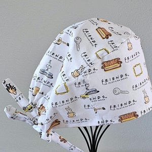 May include: White scrub hat with a Friends TV show theme. The hat has a white background with black and brown illustrations of the show's logo, a couch, a coffee cup, a key, and a guitar. The text "FRIENDS" is repeated throughout the hat.