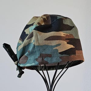 May include: Camouflage patterned scrub cap in shades of green, brown, and tan. The cap is tied at the back with a black adjustable strap. The cap is displayed on a black metal stand against a white background.