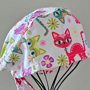 May include: A white scrub hat with a colorful cat and flower pattern. The hat has a tie at the back.