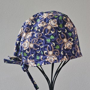 May include: A blue scrub cap with a repeating pattern of cartoon monkeys, bananas, and green leaves. The cap has a tie closure and is displayed on a black stand. The monkey design is in shades of brown and cream.