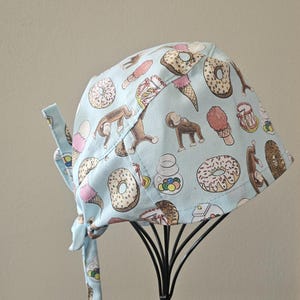 May include: A light blue scrub cap featuring a repeating pattern of brown monkeys, various donuts with sprinkles, ice cream cones, and jars of colorful candies. The cap has a tie-back closure and is constructed from fabric with a multi-colored print.
