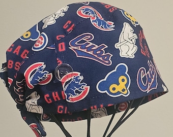 May include: A navy blue scrub cap featuring a repeating pattern of Chicago Cubs logos, bear illustrations, and the word Cubs in red and blue text. The cap includes a clear plastic button on the side for securing a face mask and tie-back closures.