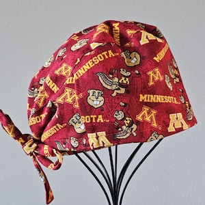 May include: A maroon and gold scrub cap featuring a repeating pattern of the University of Minnesota's "M" logo and the Goldy Gopher mascot. The cap is tied at the back with a matching fabric tie.