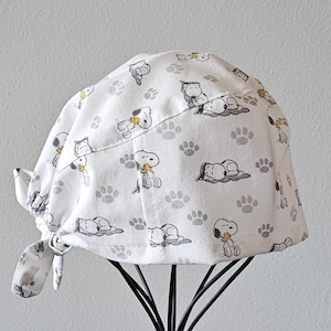 May include: White scrub hat with a tie-back closure. The hat features a repeating pattern of paw prints and cartoon images of Snoopy holding a flower.