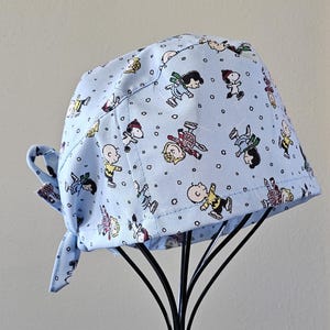 May include: A light blue surgical scrub cap featuring a repeating pattern of cartoon characters in various poses. The cap has a tie closure and is displayed on a black metal stand. The characters are in various colors, including yellow, red, and black.