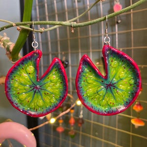 Peut inclure: Boucles d'oreilles faites à la main en forme de nénuphar. Les boucles d'oreilles sont vertes avec des bords rouges et présentent des détails de gouttelettes d'eau. Elles sont suspendues à des crochets argentés. Un accessoire unique.