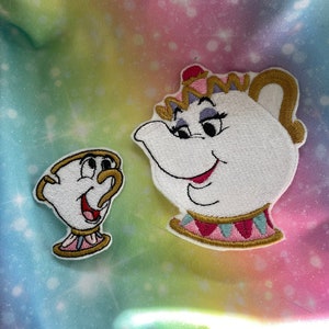Puede incluir: Dos parches bordados de personajes de la película de Disney La Bella y la Bestia. Los parches están sobre un fondo tie-dye arcoíris. El primer parche es de la Sra. Tetera, una tetera con una corona rosa y morada, una cara blanca y una boca roja. El segundo parche es de Chip, una taza de té con un asa amarilla, una cara blanca y una boca roja.