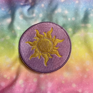 May include: A circular embroidered patch with a golden sun design inside a lavender circle. The sun has multiple pointed rays. The patch is set against a colourful, galaxy-themed background with pink, blue, and yellow shades.