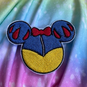 May include: Embroidered patch with a stylized design. It features blue ears, red accents, and a yellow base, resembling a character's silhouette. A red bow is centered at the top. The patch is set against a colourful, tie-dye background.