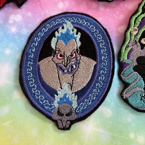 May include: A blue and white embroidered iron-on patch featuring a cartoon image of Hades from Disney's Hercules. The patch is circular and has a Greek key border.
