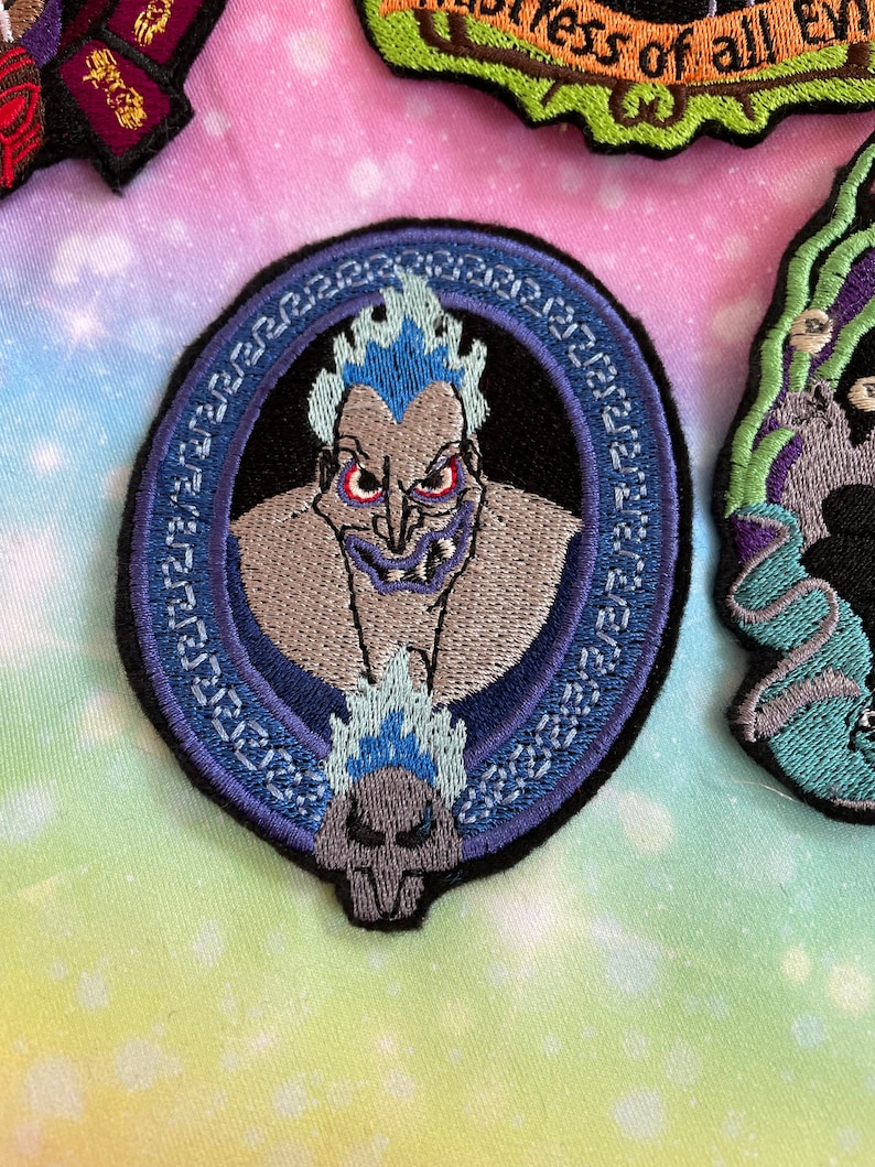 May include: A blue and white embroidered iron-on patch featuring a cartoon image of Hades from Disney's Hercules. The patch is circular and has a Greek key border.