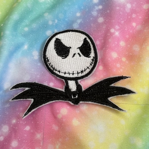 Puede incluir: Un parche bordado blanco y negro de Jack Skellington, el Rey Calabaza de Pesadilla Antes de Navidad, con una sonrisa cosida y ojos negros. El parche está sobre un fondo de tela tie-dye arcoíris.