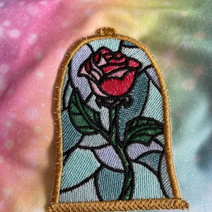 May include: A red rose with green leaves and a stem is encased in a stained glass-style frame with a gold border. The rose is in a glass dome.