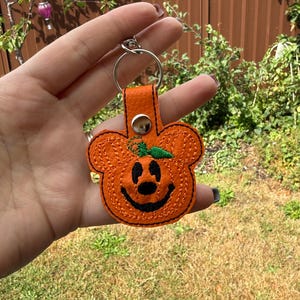 Puede incluir: Un llavero de piel sintética naranja texturizada con forma de calabaza y orejas de ratón. El diseño presenta rasgos faciales bordados en negro, un tallo bordado en verde y un cierre de botón a presión de metal plateado en una anilla.