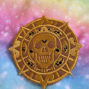 May include: A gold embroidered patch featuring a skull and crossbones design. The skull is encircled by eight symbols, each with a unique design. The patch is displayed against a rainbow tie-dye fabric background.