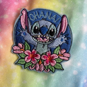May include: Blue Stitch character with big eyes and a toothy grin, saying "Ohana!" in a blue circle with pink flowers around the bottom.