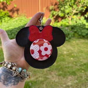 May include: Black Mickey Mouse-shaped keyring with a red glitter bow and a round button. The button features a white polka dot and flower design on a red background. The keyring is attached to a silver metal ring.