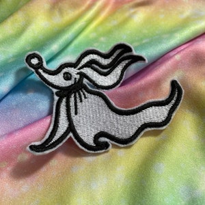 May include: White and black embroidered iron-on patch of Zero, the ghost dog from the Nightmare Before Christmas film. The patch is on a rainbow tie-dye fabric.